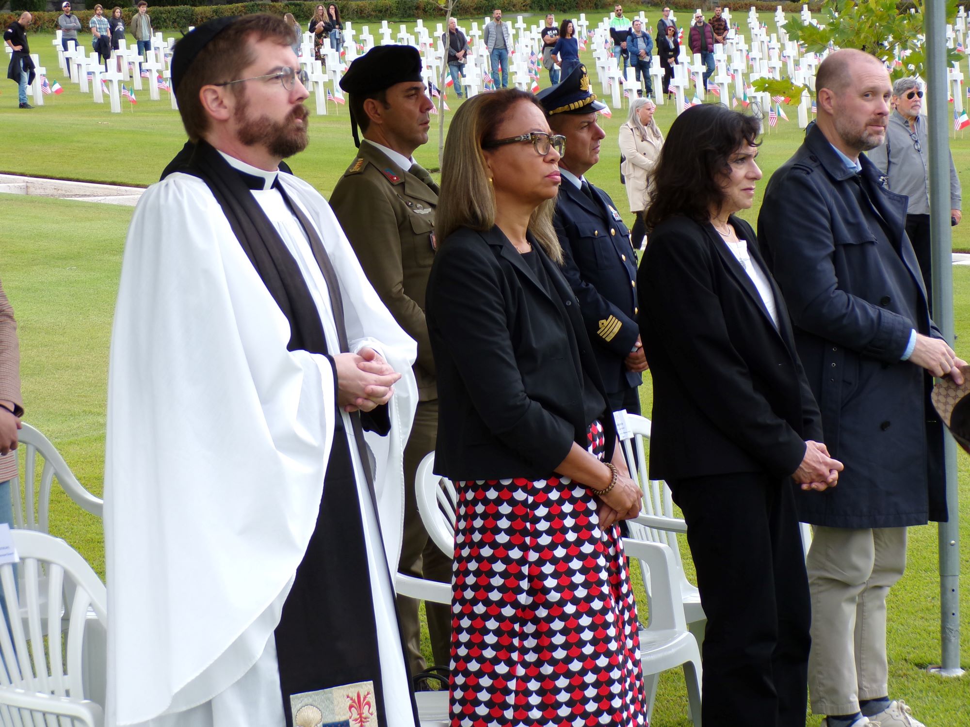 Ceremony participants stand under a tent at the cemetery. In the background others stand beside headstones.