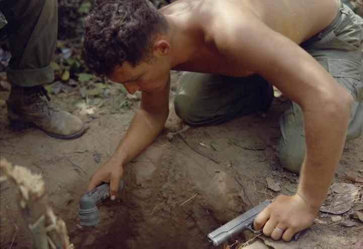 A shirtless man peers down into a hole with a flashlight.