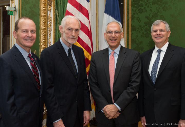 Four men in suits stand in front of an American and French flag. 