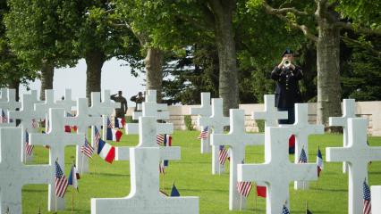 A bugler stands amidst headstones, each marked with an American and French flag. 