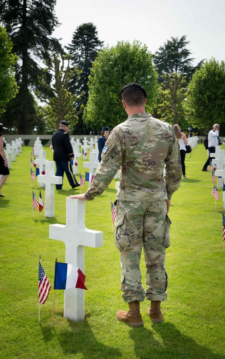A soldier in fatigues stands next to a headstone. 